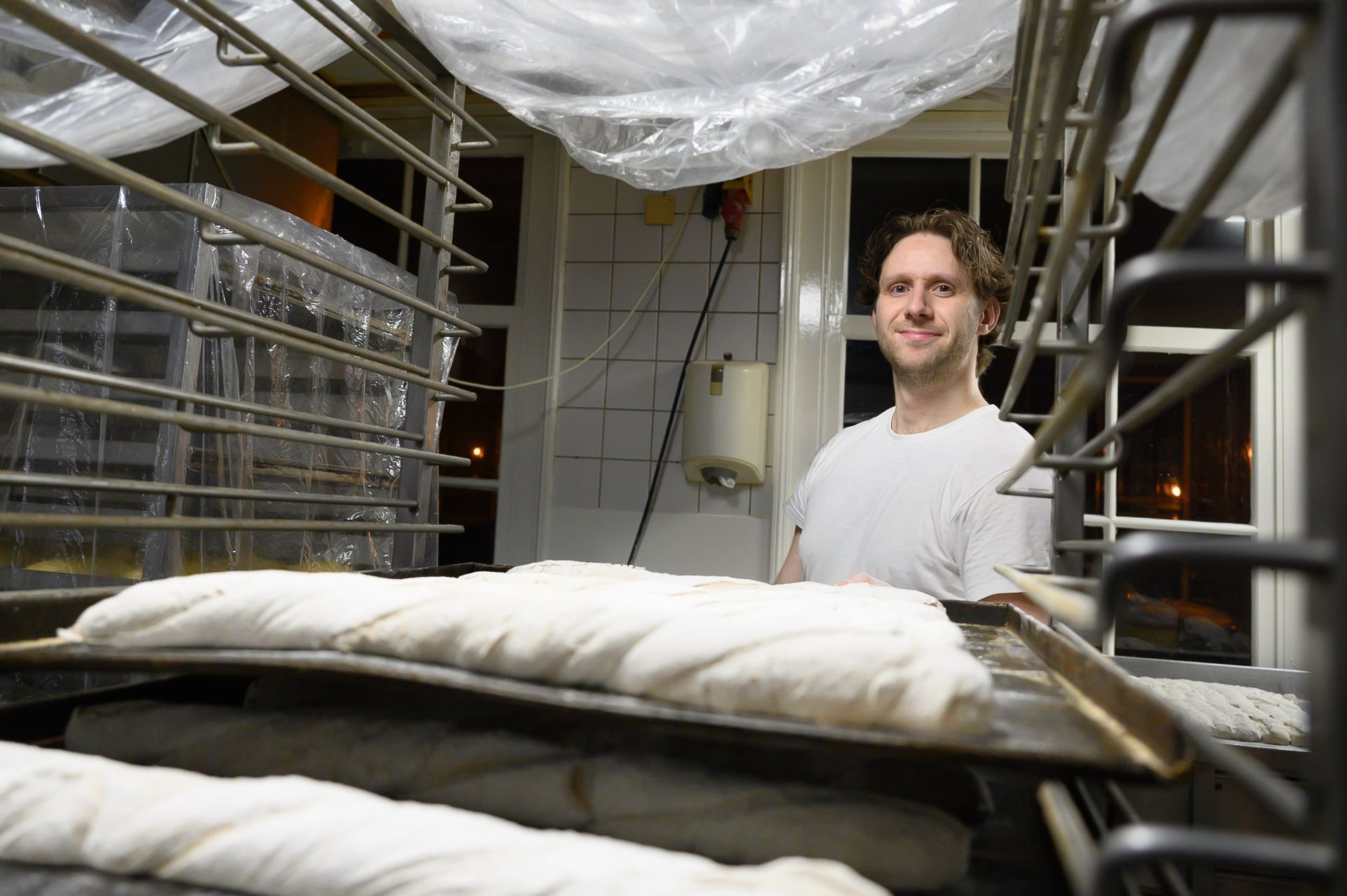 Een bakker glimlacht in een professionele keuken naast schalen met rijsdeeg onder plasticfolie.