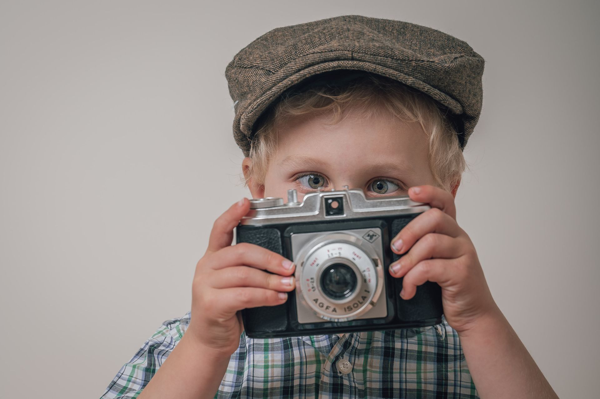 Kind in een geruit shirt en pet houdt een vintage camera voor zijn gezicht tegen een effen achtergrond.
