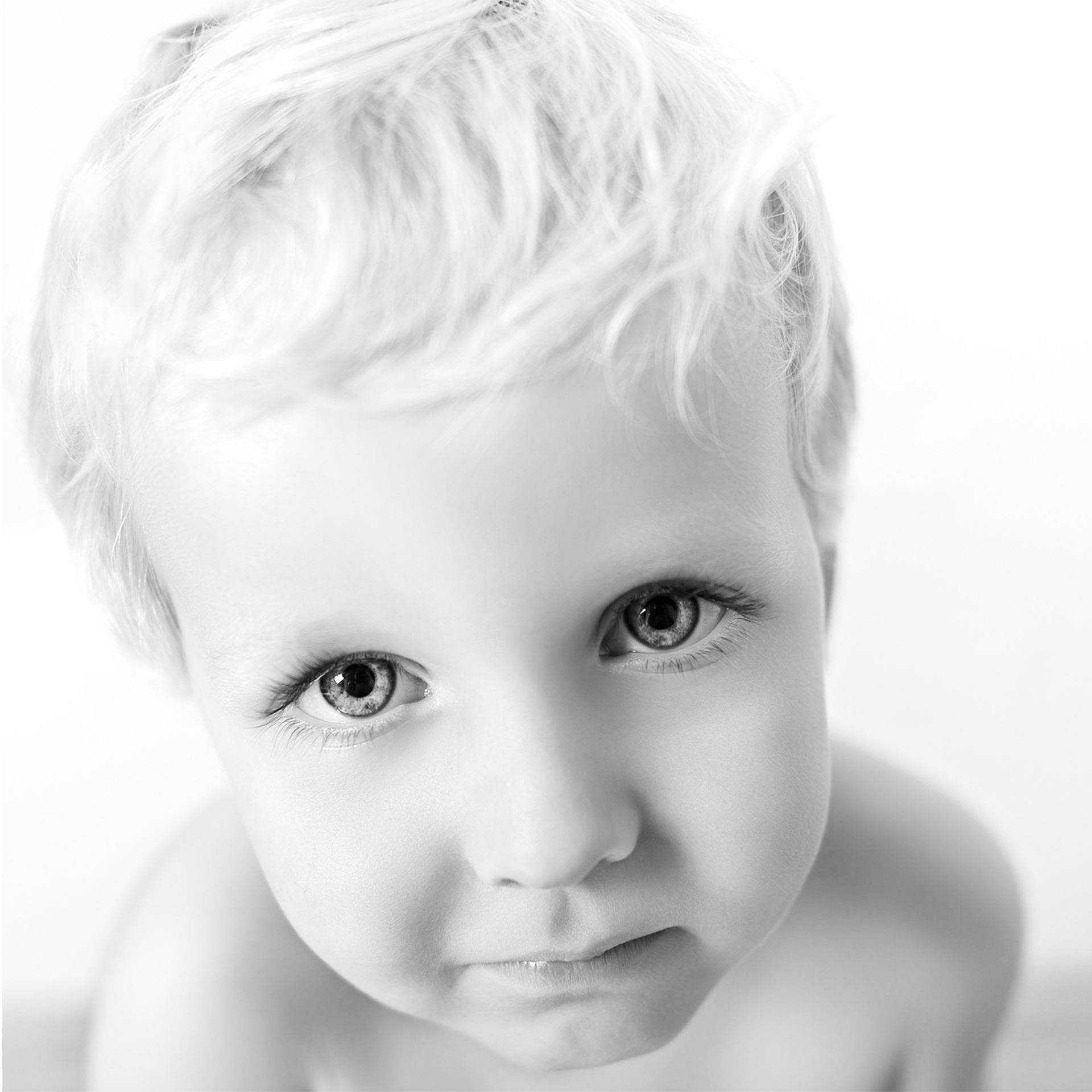 Close-up van een blond kind dat met een kalme uitdrukking omhoog kijkt tegen een witte achtergrond.