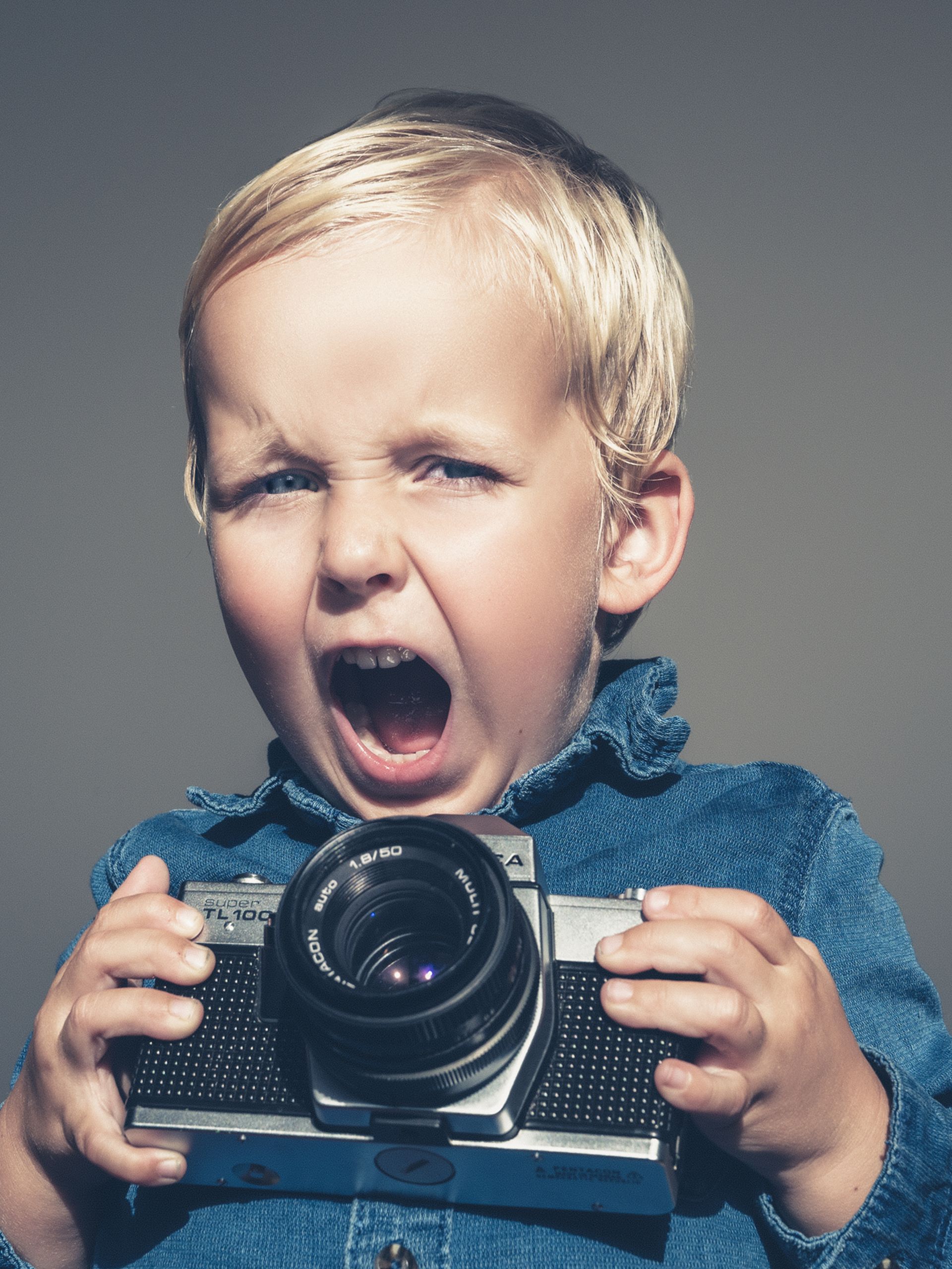 Een kind gaapt terwijl het een vintage camera vasthoudt tegen een grijze achtergrond.