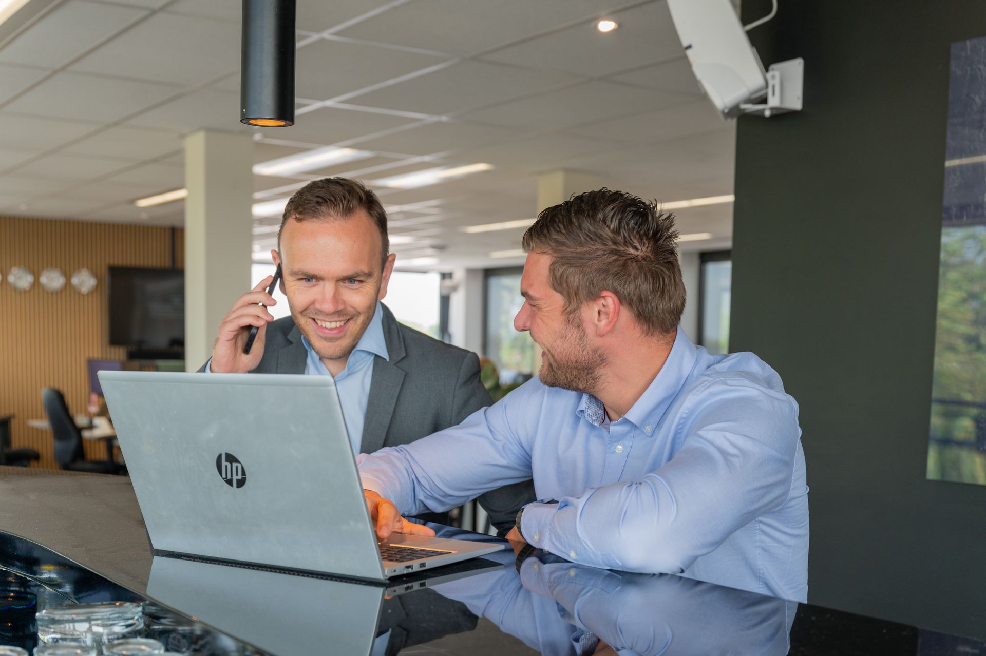 Twee collega's glimlachen en praten via een laptop in een moderne kantoorlounge.