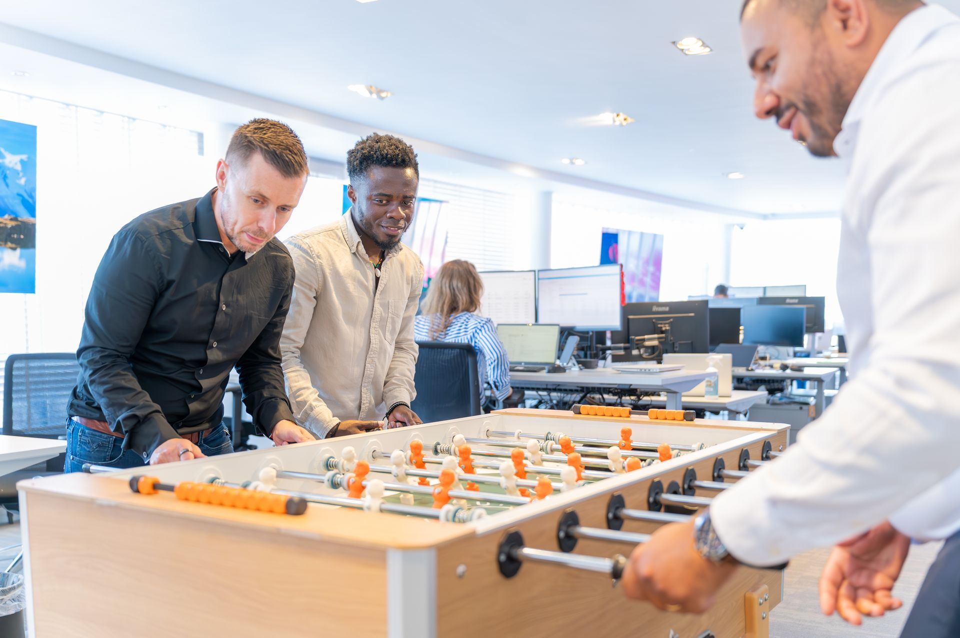 Mensen spelen tafelvoetbal in een lichte kantoorlounge met computers op de achtergrond.