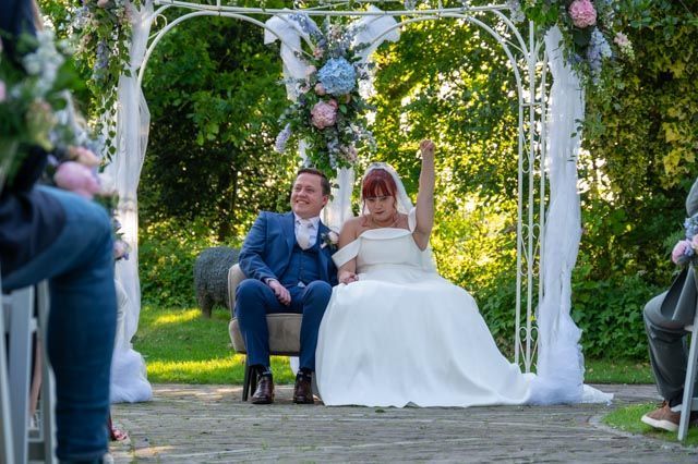 Bruid en bruidegroom onder een bloemenboog, terwijl ze hun handen omhoog steken tijdens een huwelijksceremonie in de buitenlucht.