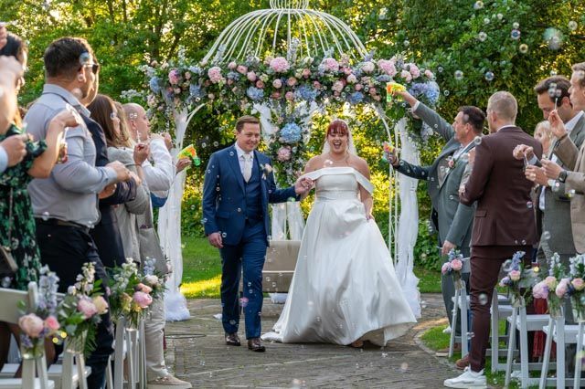 Bruid en bruidegom lopen hand in hand door een met bloemen versierde tuinpad onder een bloemenboog tijdens een huwelijksceremonie.