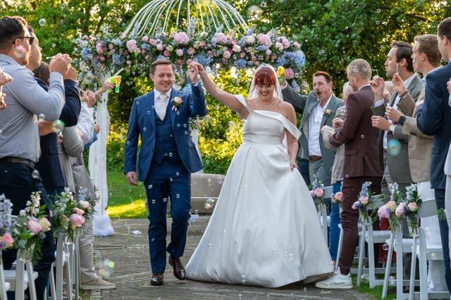 Bruid en bruidegroom lopen naar het altaar tijdens een buitenbruiloft, onder luid applaus van de gasten onder een bloemenboog.