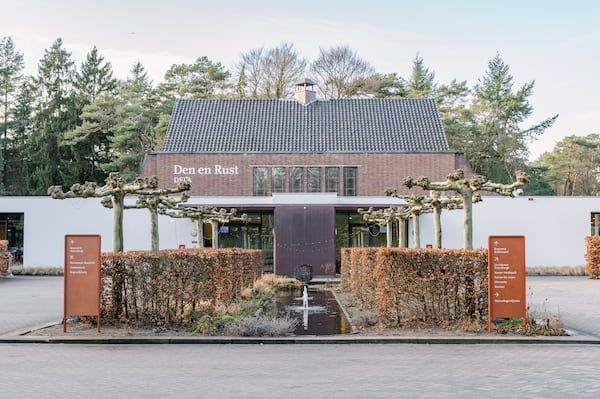 Een klein bakstenen gebouw met een donker dak, een heg aan de voorkant en bomen erachter, onder een bewolkte hemel.
