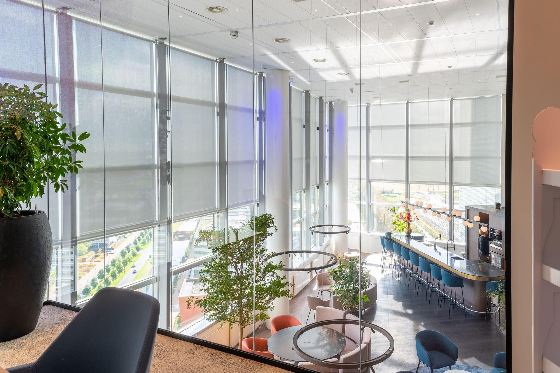 Licht en ruim kantooratrium met grote ramen, glazen balustrades, planten en een zithoek met uitzicht op de bureaus beneden.