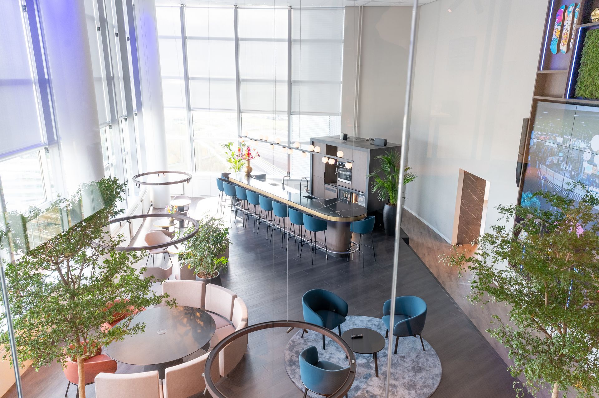 Lichte café-lounge met ronde tafels, turquoise stoelen, planten en een lange bar bij grote ramen.
