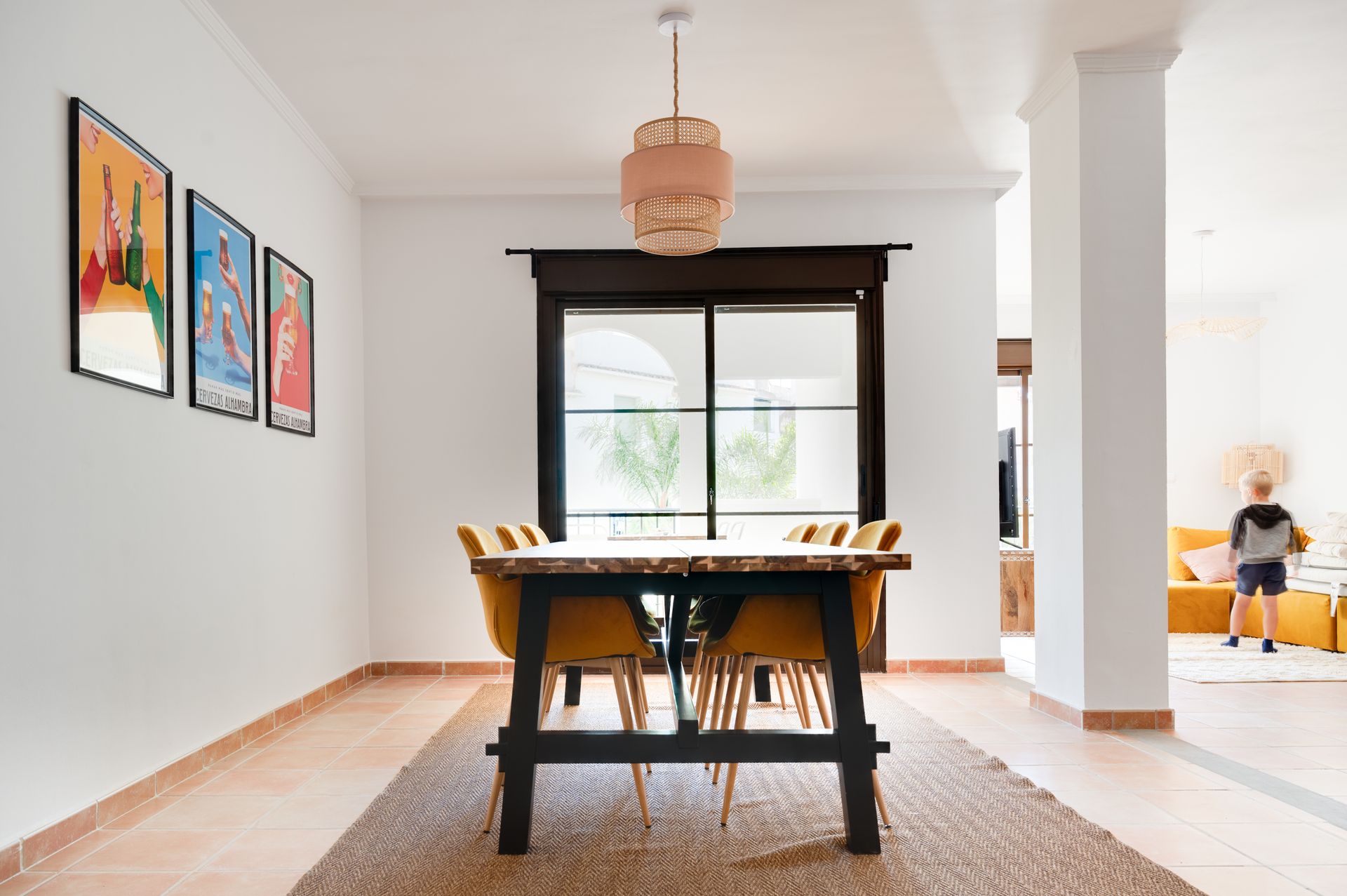 Lichte eetkamer met houten tafel, stoelen, een geweven hanglamp en ingelijste kunst bij een raam en een open deur.