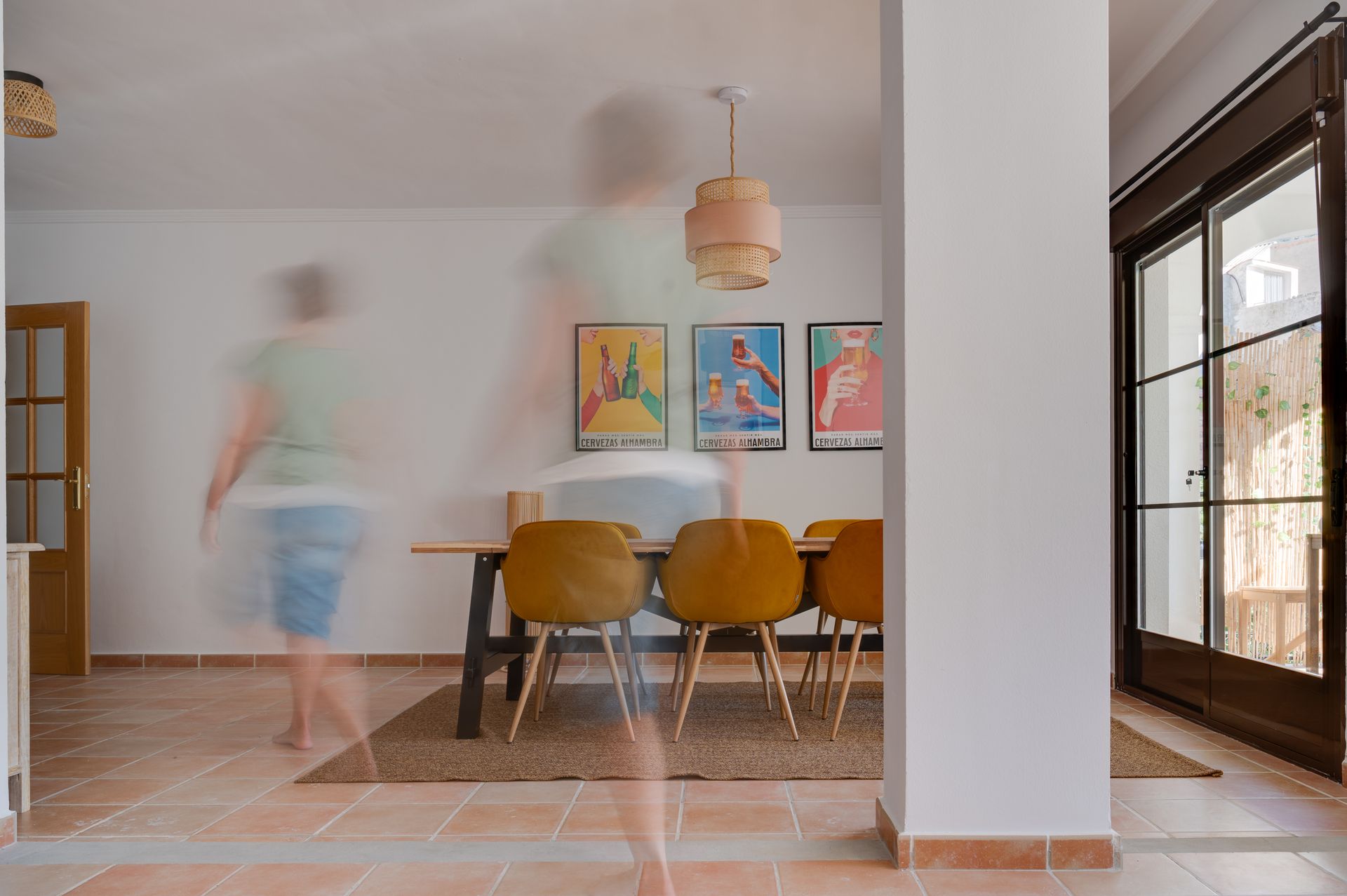 Lichte eetkamer met houten tafel, oranje stoelen, kleurrijke muurdecoratie en een wazige figuur die voorbijloopt.