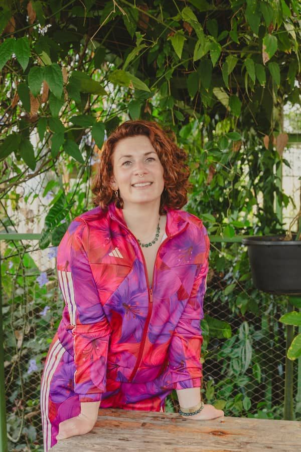 Vrouw in een felroze-paarse blouse die tegen een reling leunt onder weelderig groen.