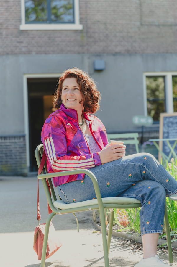 Een vrouw zit buiten op een stoel, gekleed in een felroze jas en spijkerbroek, voor een gebouw.