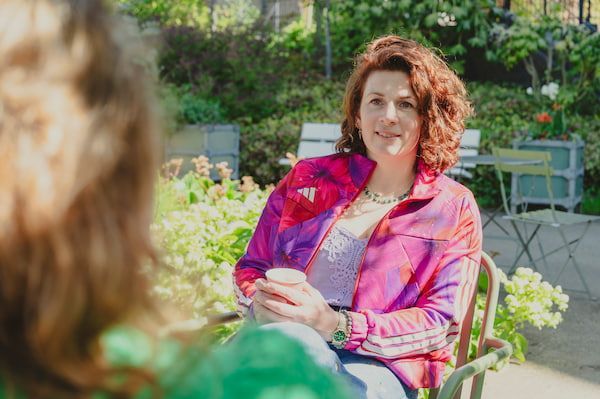 Vrouw in een roze jas zit buiten en praat met iemand op de voorgrond.