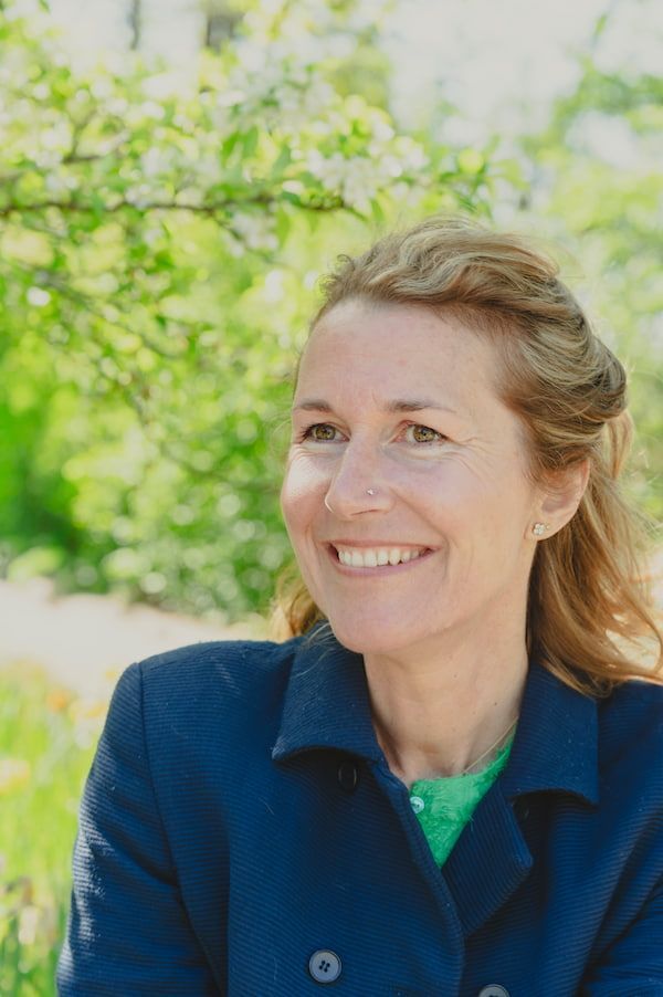 Lachende vrouw in een blauwe jas buiten met groen gebladerte op de achtergrond.