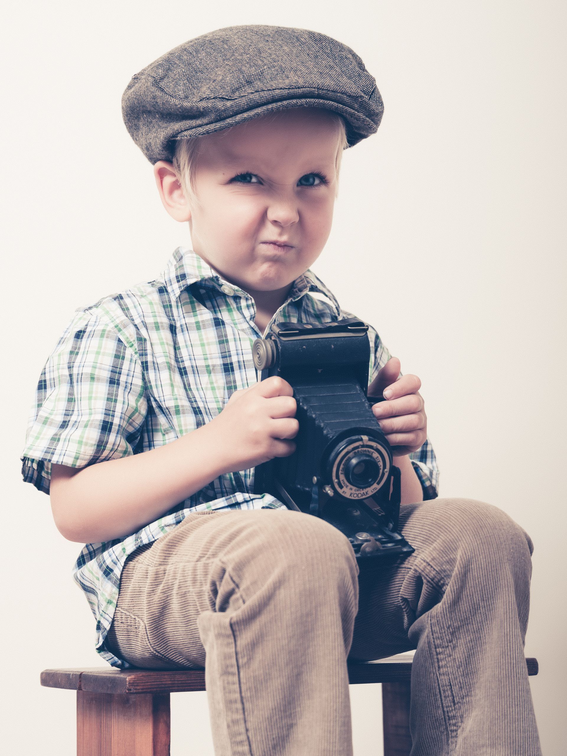 Jong kind met een platte pet, een vintage camera in de hand en een strenge uitdrukking op zijn gezicht.