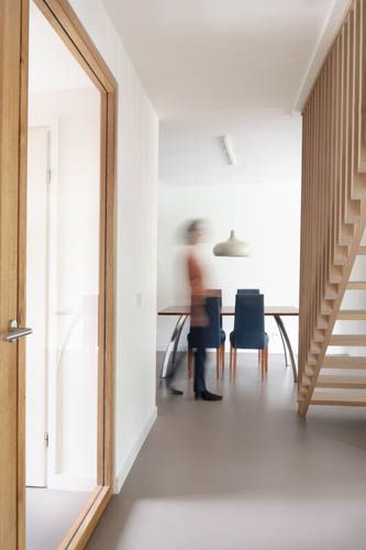 Moderne hal met houten trap, witte muren en een wazig figuur dat bij een eettafel staat.