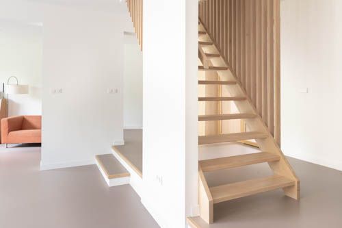 Licht, minimalistisch interieur met een trap, witte muren en lichte houten treden naast een zithoek.
