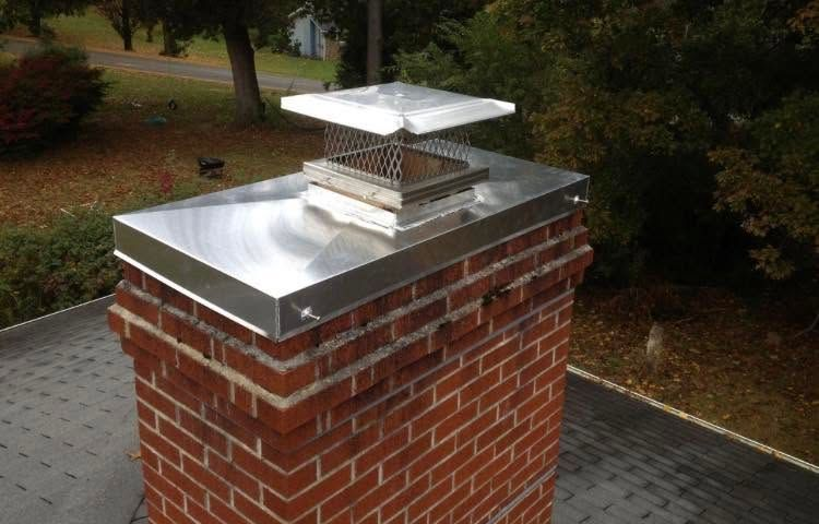 Brick chimney with a silver metal cap on a shingled roof, surrounded by trees.
