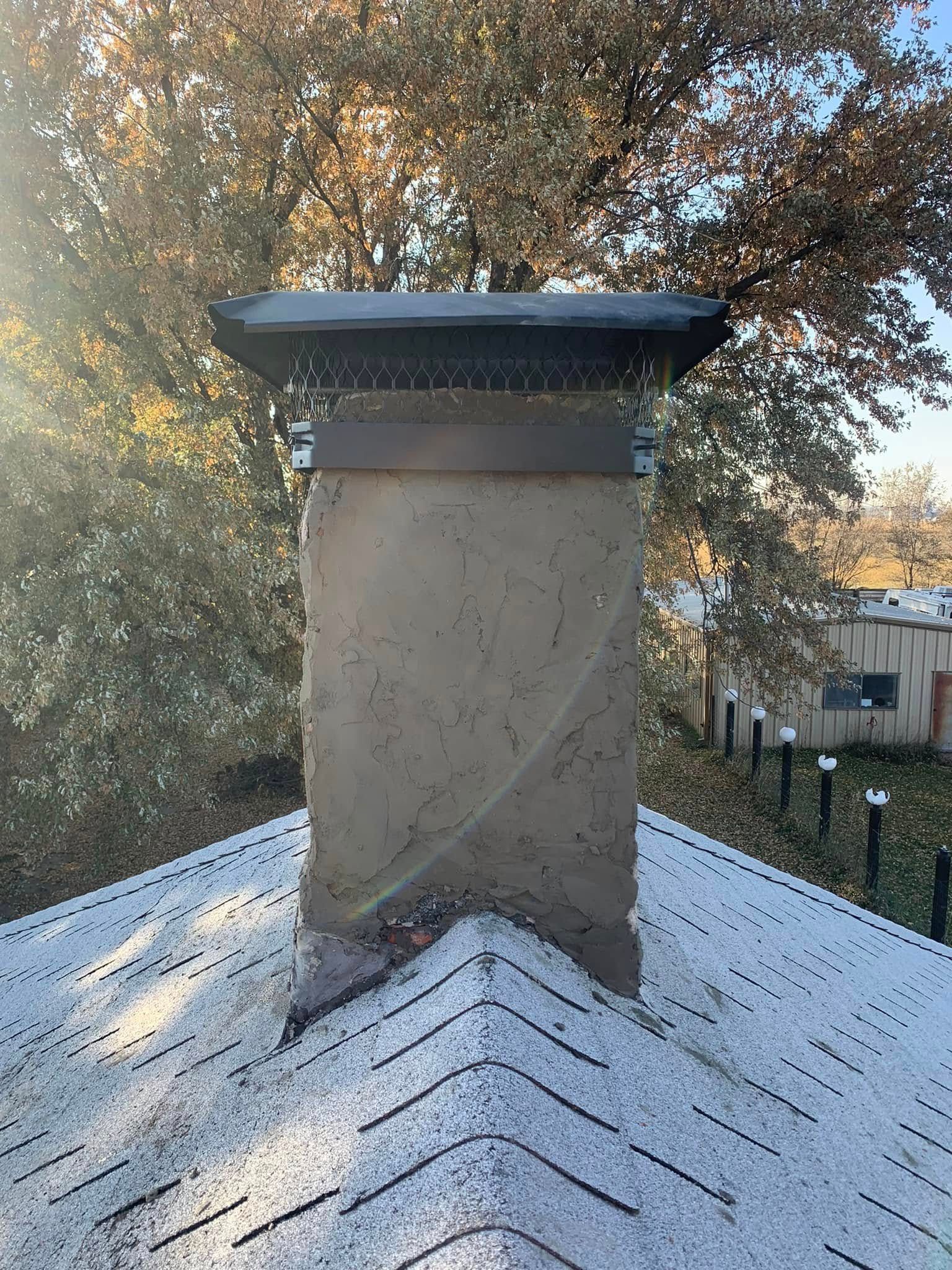 A rooftop chimney with a black cap and white shingles, framed by trees in autumn light.
