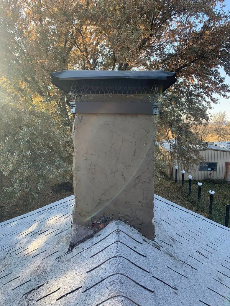 A rooftop chimney with a black cap and white shingles, framed by trees in autumn light.