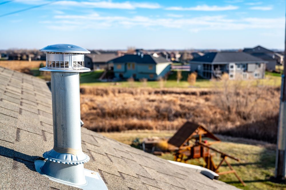 Metal roof vent on a shingled rooftop overlooking suburban houses under a blue sky.