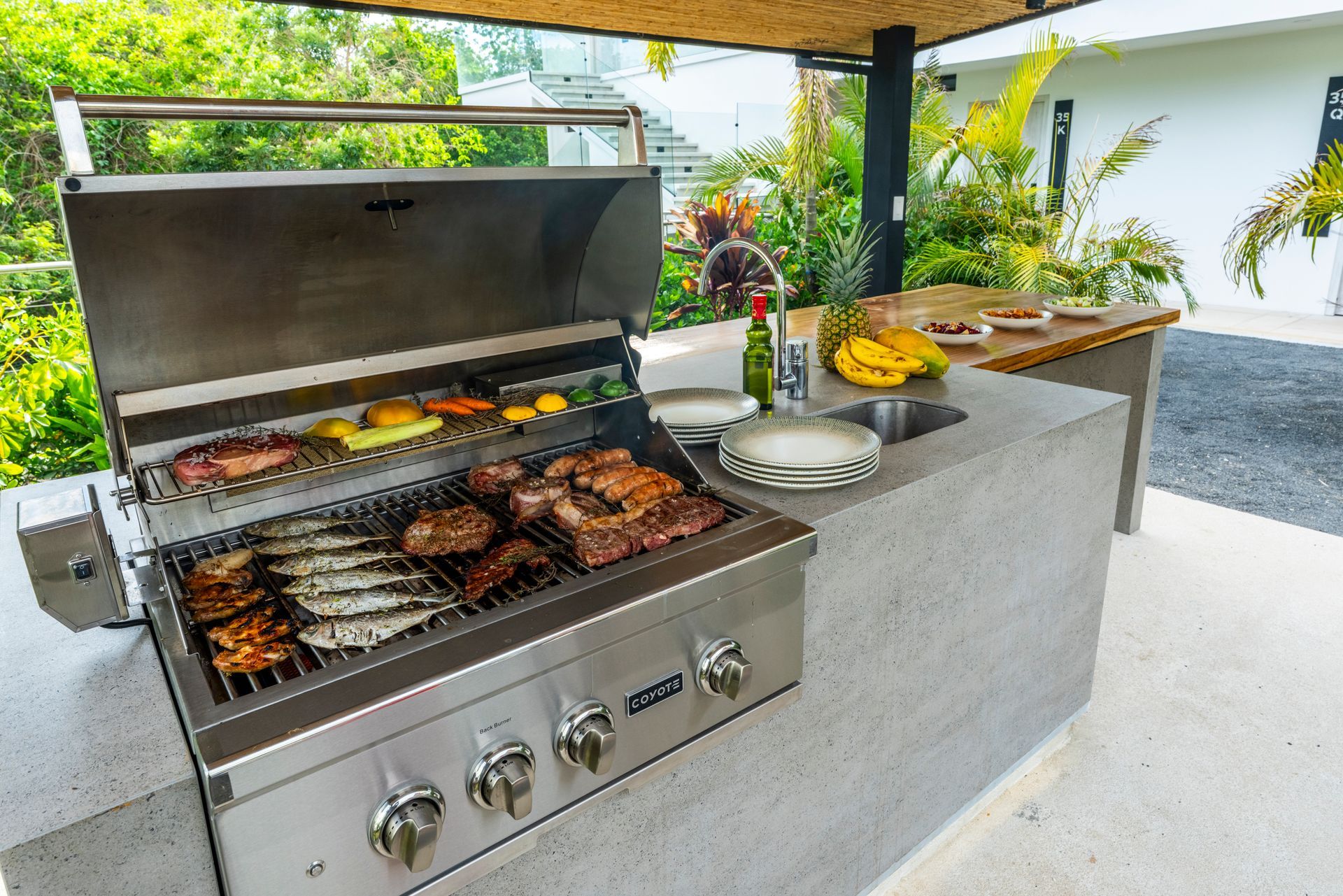 Outdoor grill with sizzling meat and vegetables on a patio countertop, with fruit and serving dishes nearby