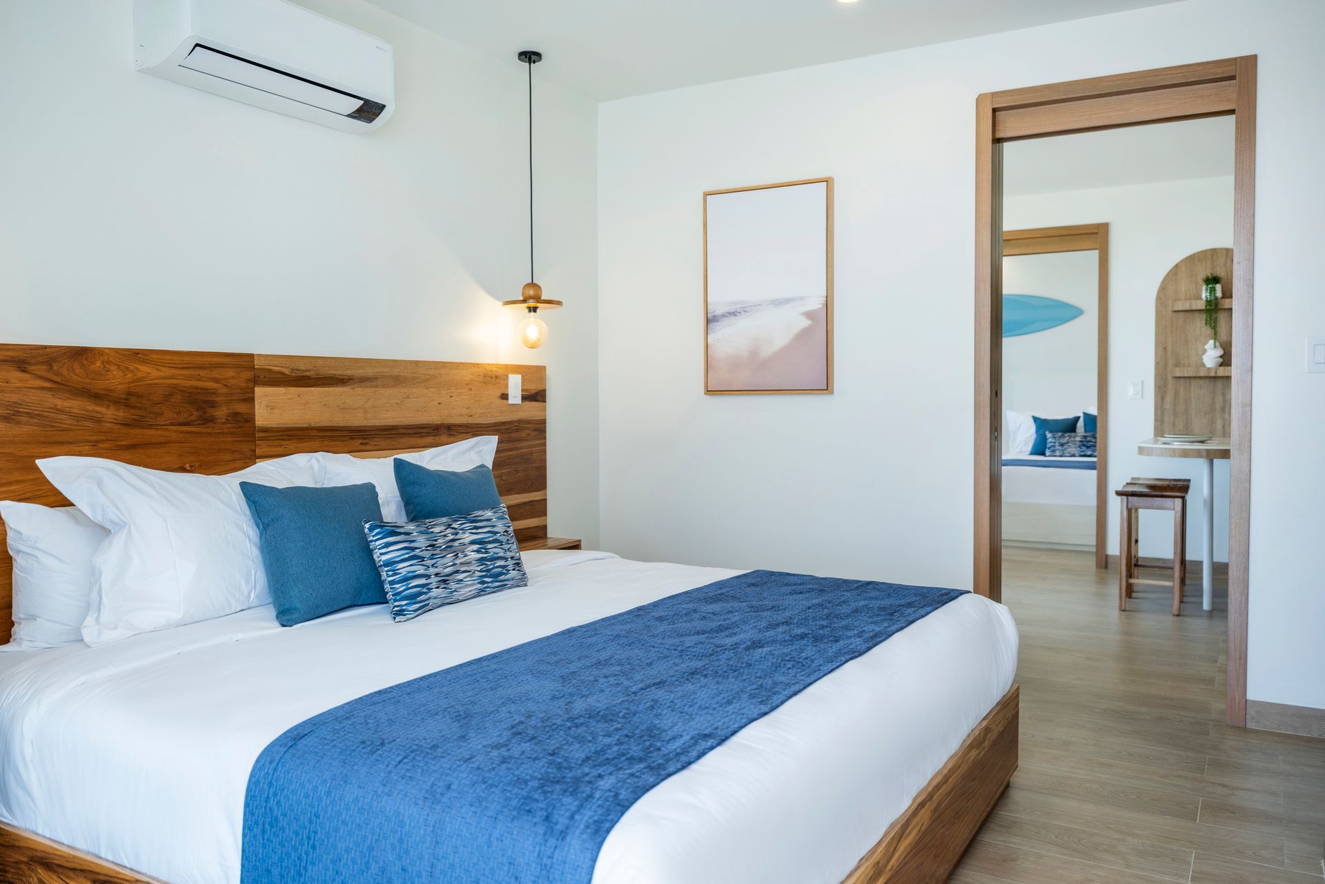 Modern bedroom with white bedding, blue accents, wood headboard, and an open doorway to a seating area