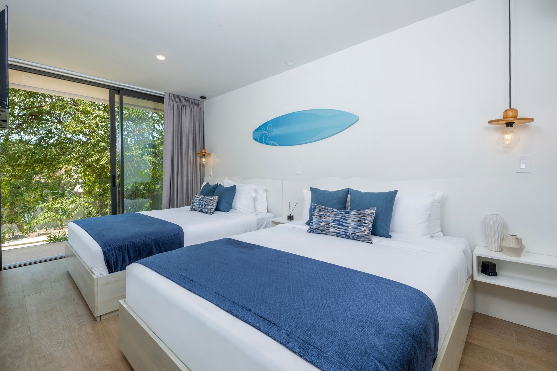 Modern bedroom with two white beds, blue bedding, wall art, and large windows overlooking greenery