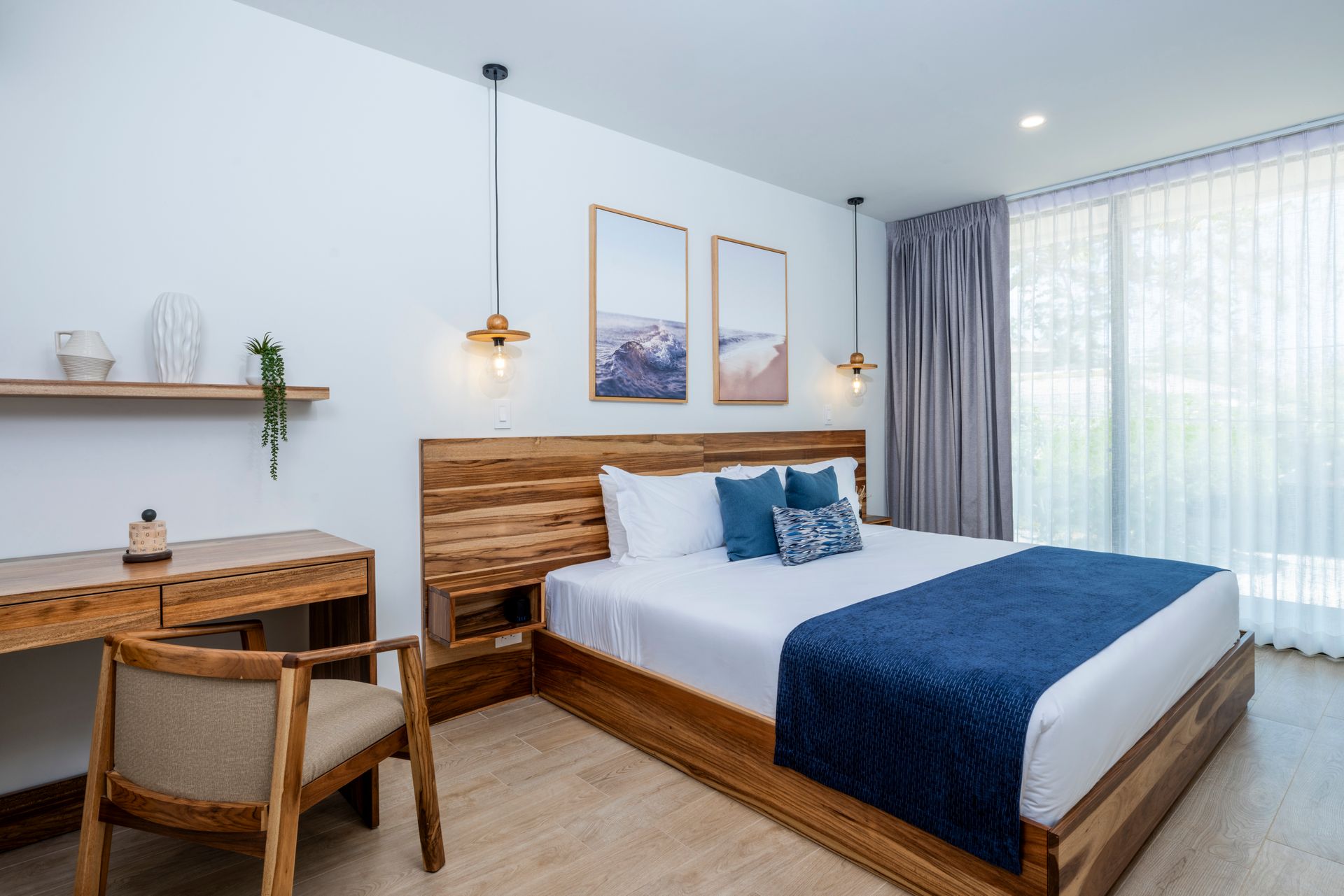Bright bedroom with a wooden bed, blue bedding, desk, wall art, and large curtained window