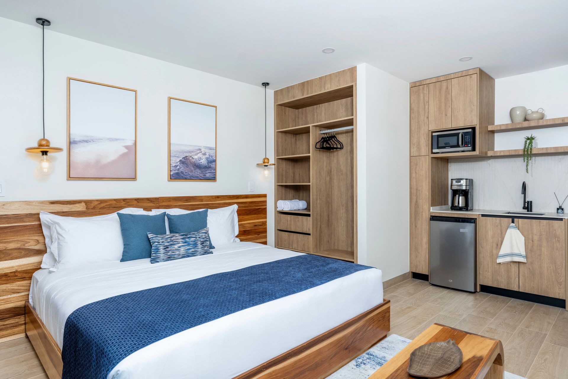 Modern bedroom with a large bed, blue bedding, framed art, and a small kitchenette with fridge and shelves