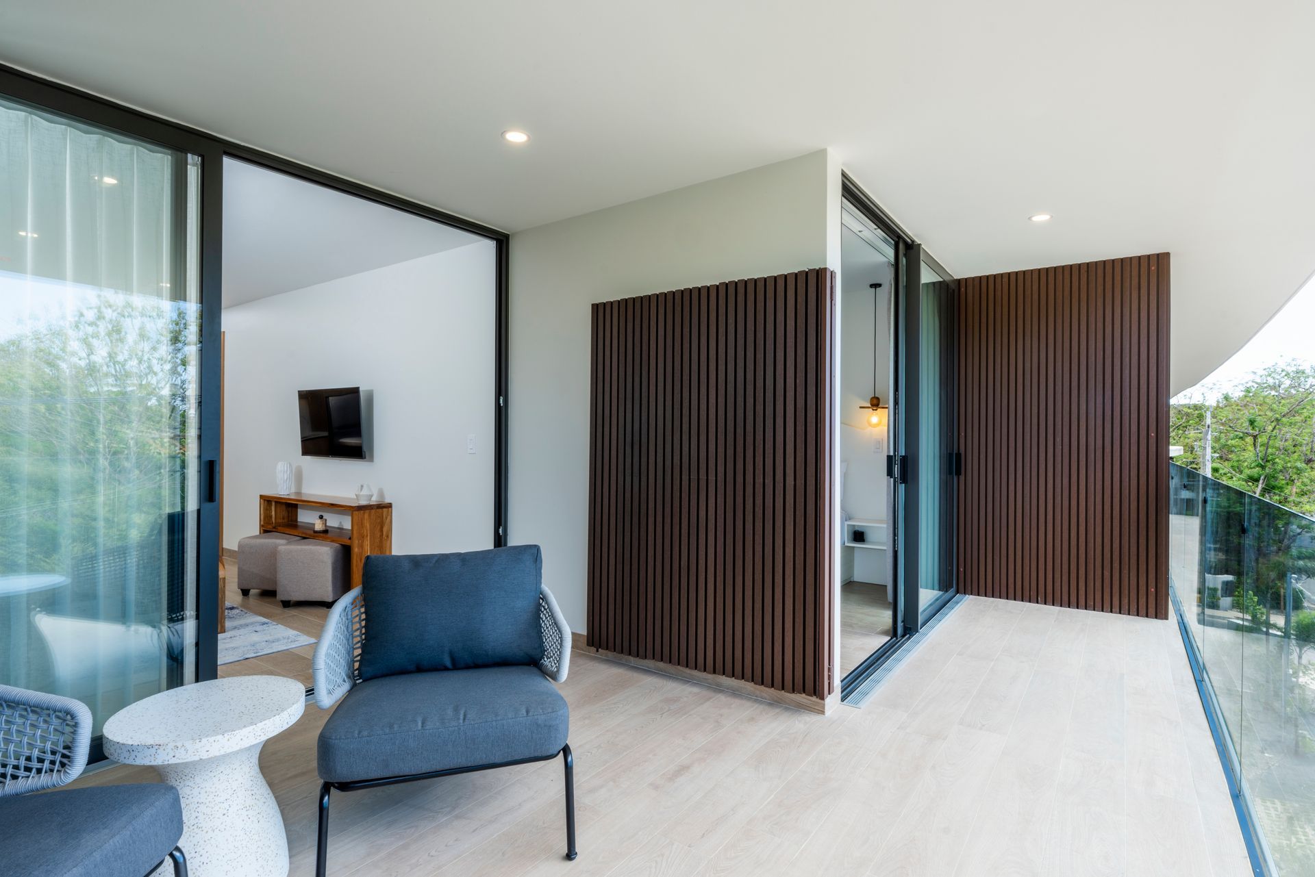 Modern outdoor patio with blue chairs, white tables, and wood-slatted walls beside a glass-walled home