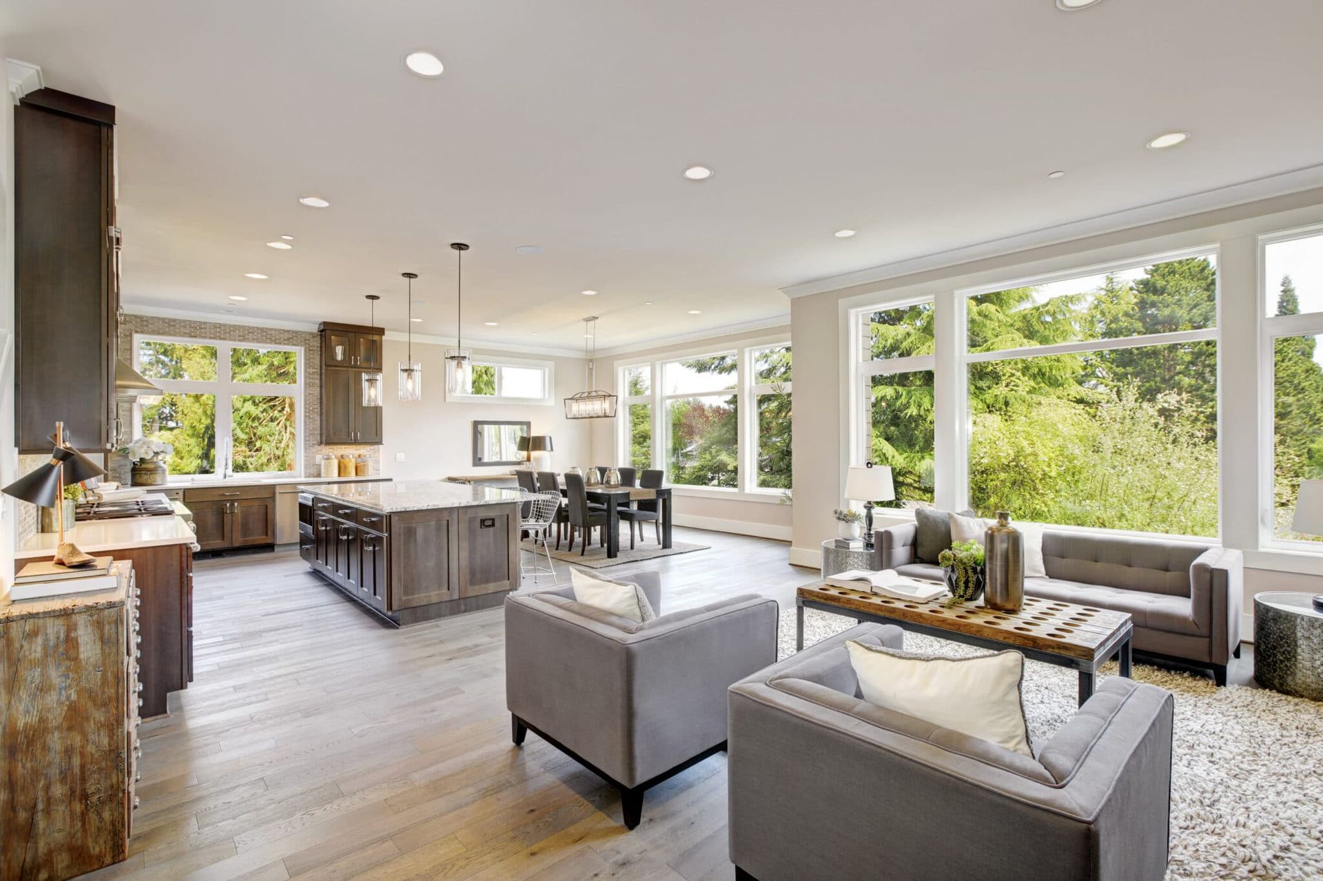 Spacious open-concept living area with kitchen, dining area, and living room. Large windows offer a view of trees.