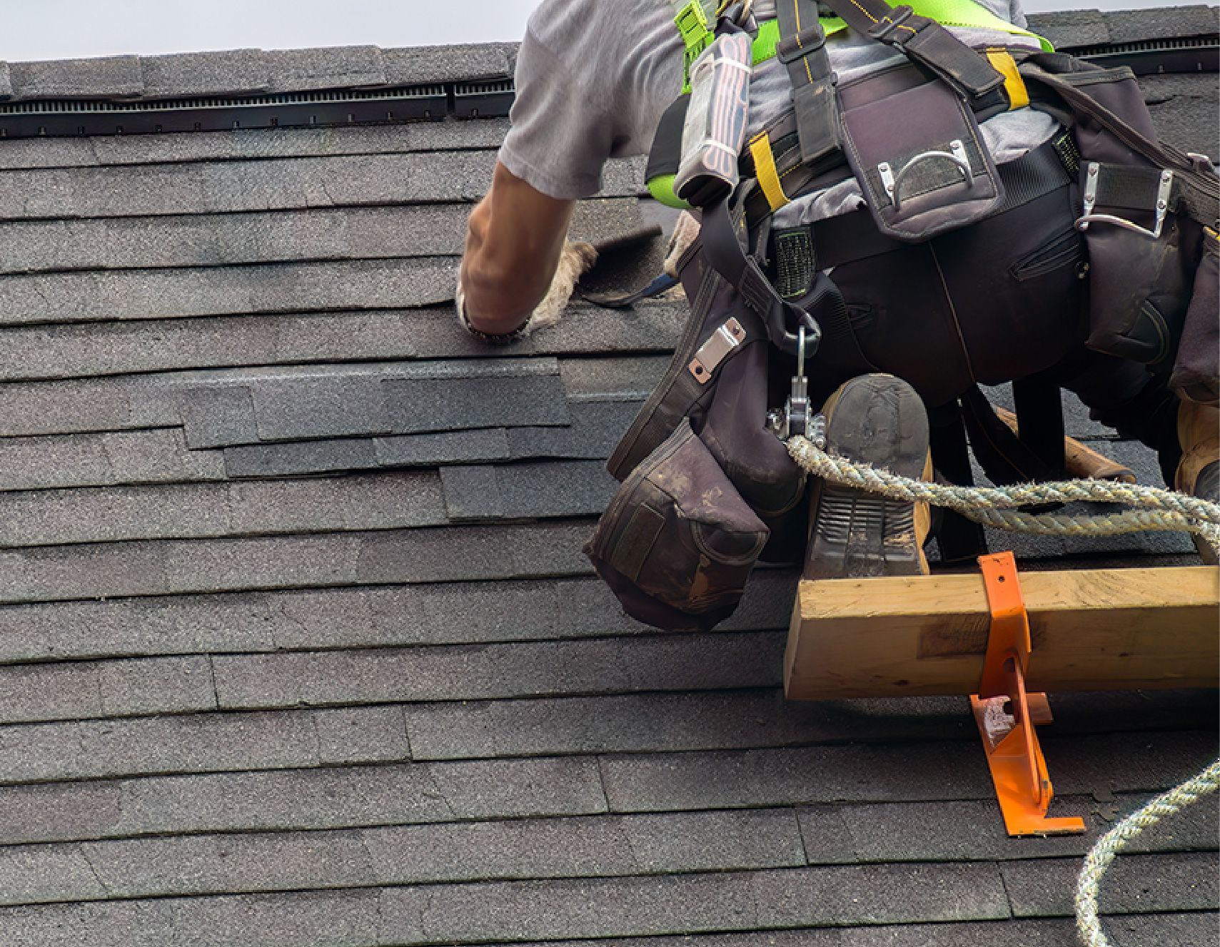 Roofer on a dark shingle roof, wearing a safety harness, repairing damaged shingles.