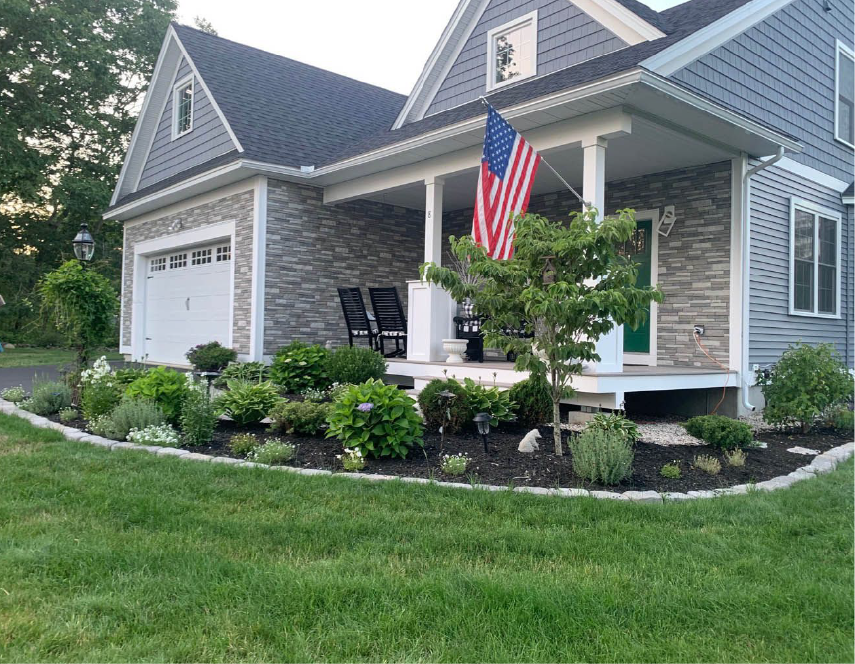 Front of a house with a porch, American flag, and manicured garden with a stone facade.