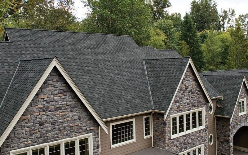 Dark gray shingle roof on a house with stone siding and multiple gables, surrounded by trees.