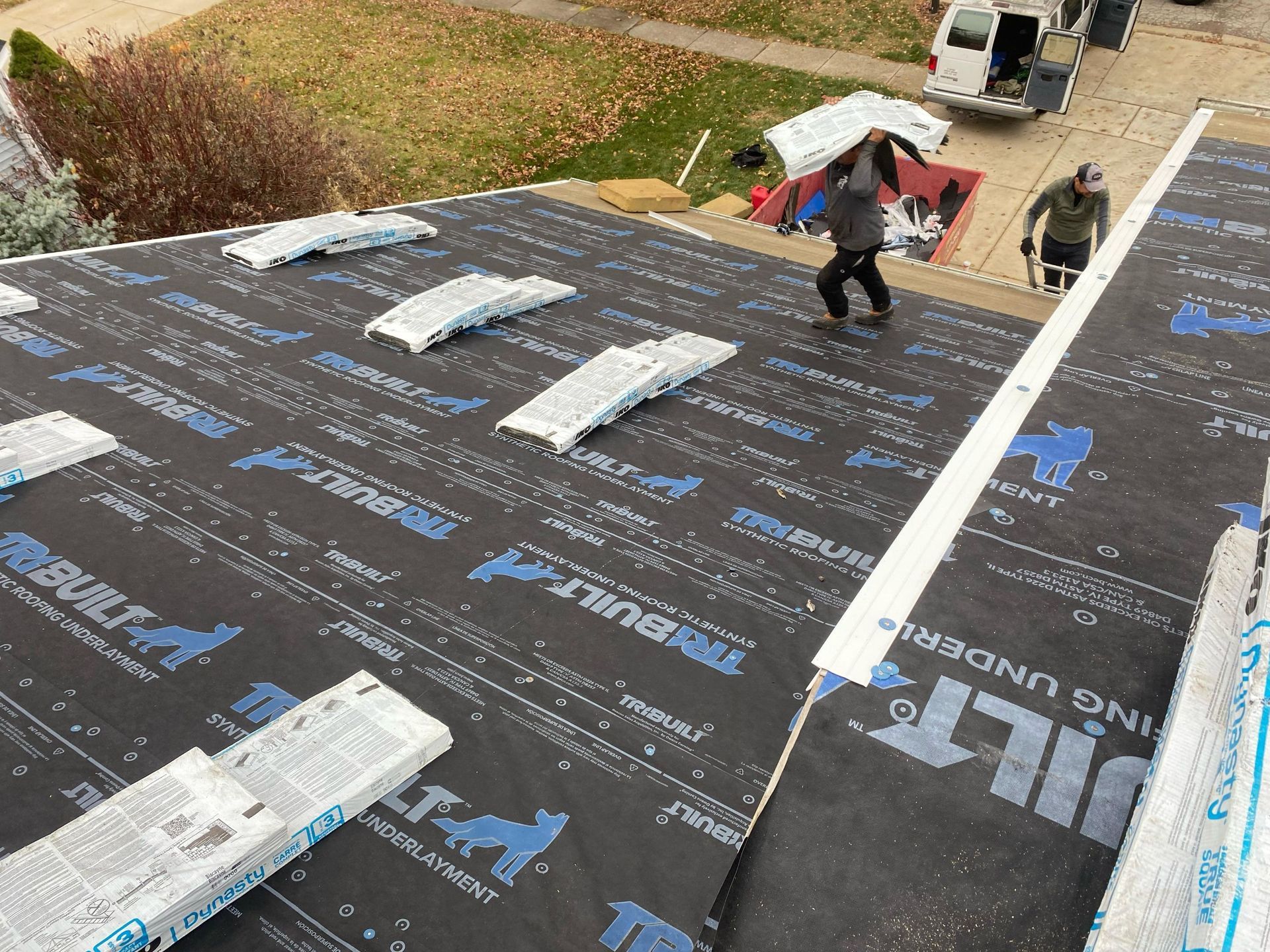 Roofer using a nail gun to install gray shingles on a rooftop. Worker wearing safety vest and gloves.