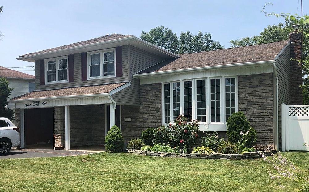 Two-story brick house with bay window, carport, and brown roof under a blue sky.