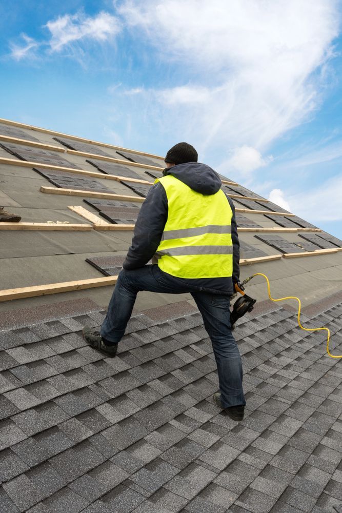 Roofer wearing a yellow safety vest on a roof, installing shingles against a blue sky.