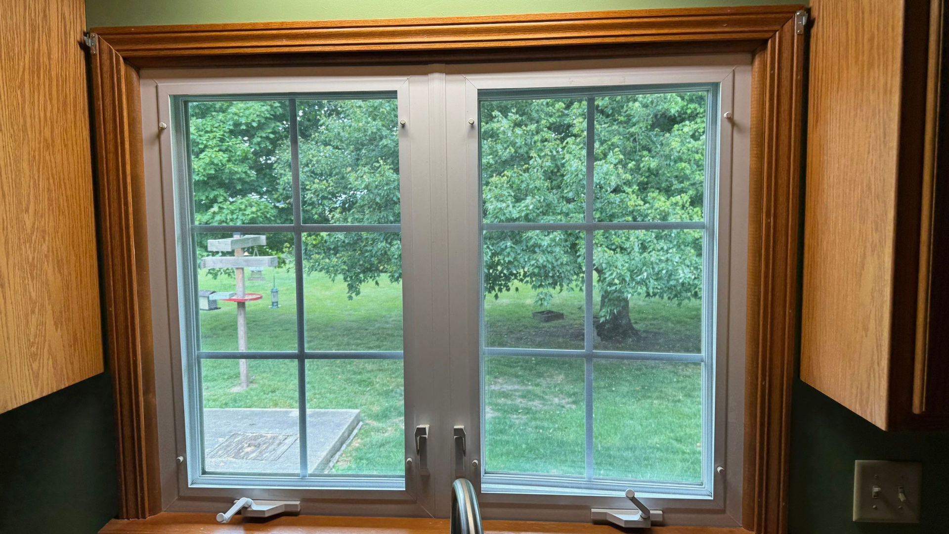 Wooden-framed window overlooking a green yard and trees, with a sink faucet at the bottom foreground