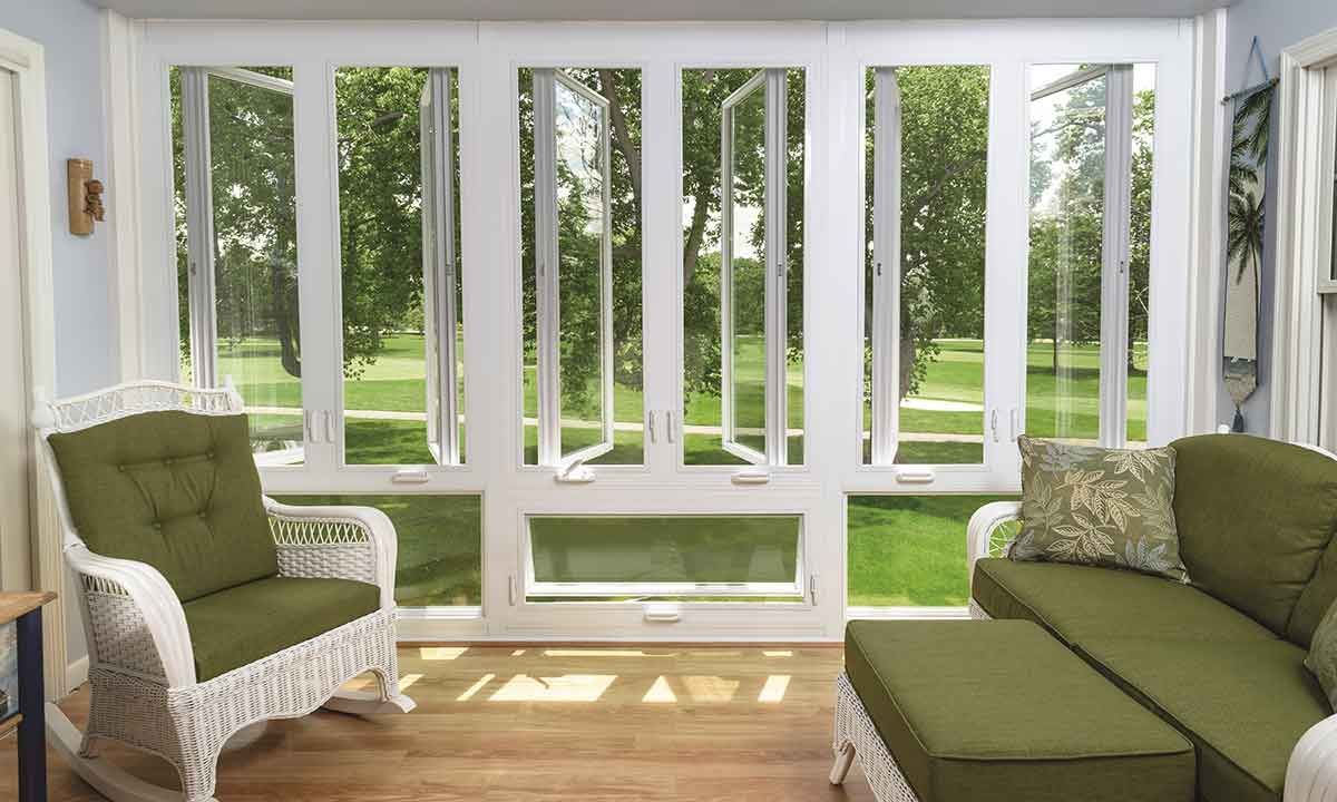 Sunroom with white wicker furniture and green cushions, overlooking a green yard with trees.