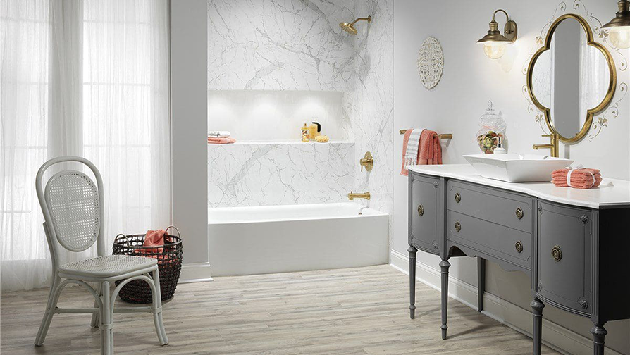 Bathroom with marble shower, antique vanity, gold fixtures, and a white chair.