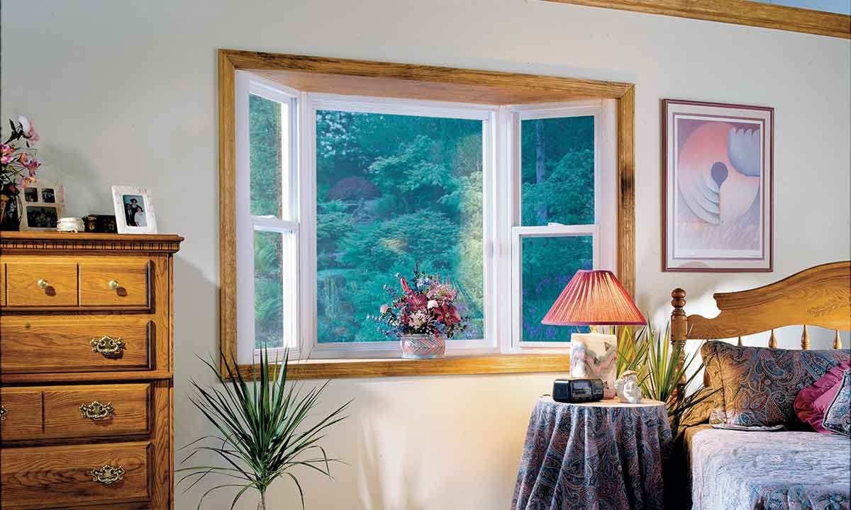 Bedroom with a bay window overlooking a forest; wooden furniture, artwork, and a table lamp.