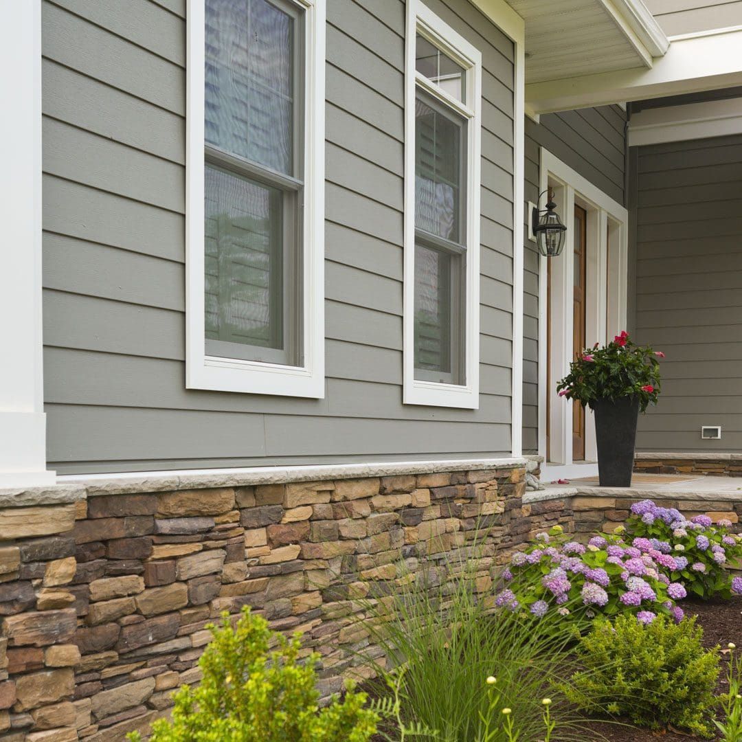 House exterior with gray siding, stone base, white-framed windows, and a doorway. Landscaping includes flowers.