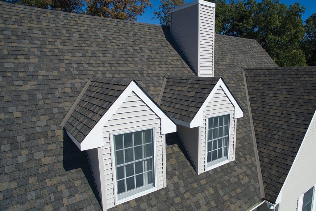 Asphalt shingle roof with two dormer windows and a chimney against a blue sky.