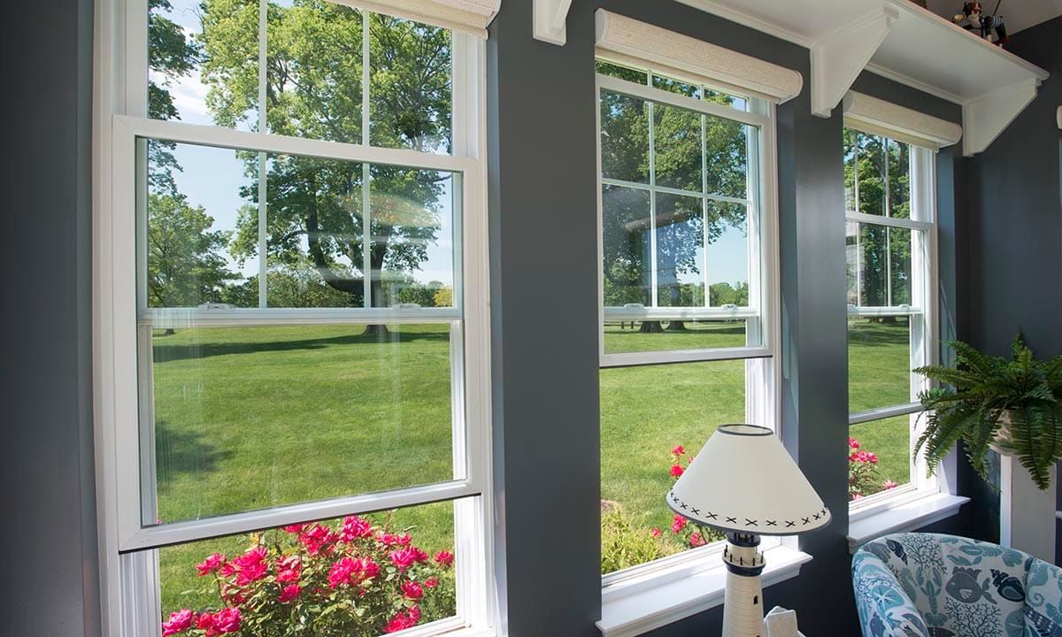 Three white-framed windows overlooking a green lawn with trees, flowers, and a table lamp.