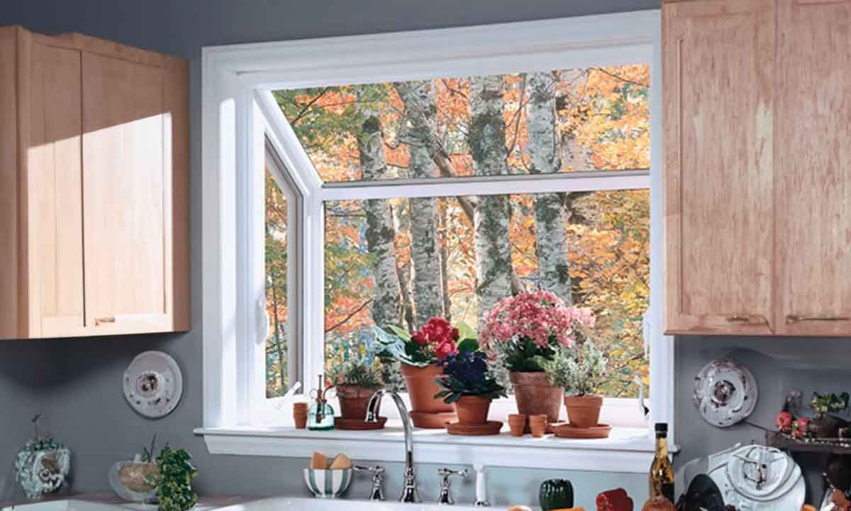 Kitchen window open to autumn trees; plants on the windowsill, wooden cabinets on either side.
