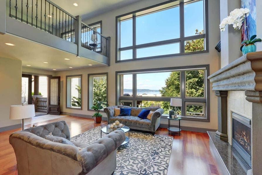 Spacious living room with large windows, fireplace, two-story ceiling, and a view of trees and water.