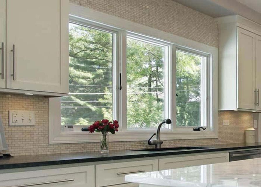 Kitchen with white cabinets, dark countertop, sink, and triple window overlooking trees.
