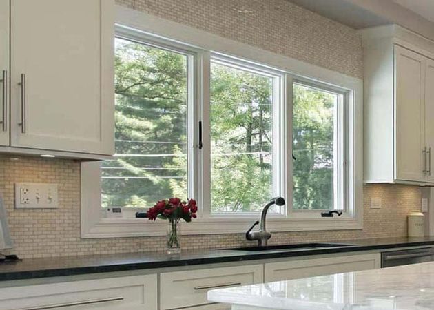 Kitchen with white cabinets, dark countertop, sink, and triple window overlooking trees.