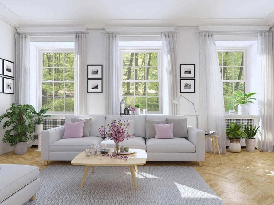 Bright living room with white sofa, windows, and plants. Light wood floor, gray rug, and pink pillows.